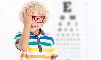 blonder Junge mit roter Brille hält sich ein Auge zu