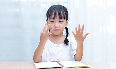 Kleines Mädchen sitzt vor einem Schulbuch und zaehlt mit den fingern