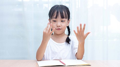 Kleines Mädchen sitzt vor einem Schulbuch und zaehlt mit den fingern