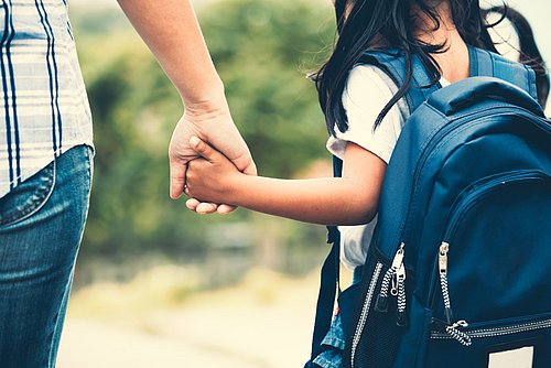 Kind mit Rucksack hält die Hand eines Erwachsenen