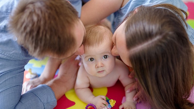 Baby_liegt_auf_einem_weichen_boden_und_wird_von_Mutter_und_Vater_gekuesst