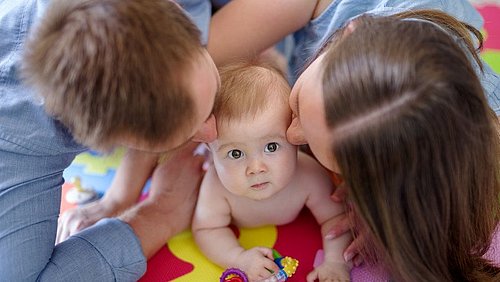 Baby_liegt_auf_einem_weichen_boden_und_wird_von_Mutter_und_Vater_gekuesst