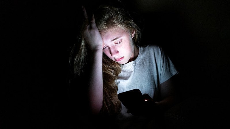 Umgeben_von_Dunkelheit_schaut_ein_Mädchen_mit_starkem_Unbehagen_auf beleuchtetes_Handy_Display