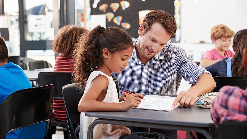 Junger_Lehrer_hilft_Mädchen_mit_Aufgabe_im_Klassenzimmer