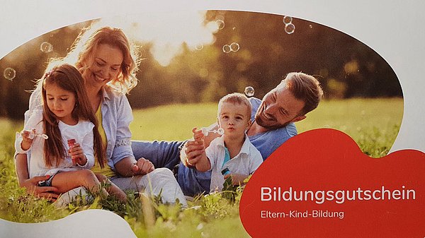 Das Bild zeigt eine Familie, die auf einer Wiese sitzt und Seifenblasen macht. In der rechten unteren Ecke findet sich ein Schriftzug mit dem Titel &quot;Bildungsgutschein&quot;
