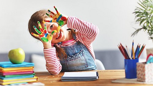 Kind sitzt mit bunt bemalten Händen an einem Schreibtisch mit Büchern und Stiften