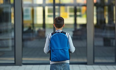 Kind steht mit Rucksack for den großen Glasstüren einer Schule